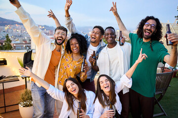 Group of diverse excited friends celebrating with beers at barbecue party in attic. Portrait of young people posing raising arms on terrace in evening in open air. Cheerful colleagues enjoying time.