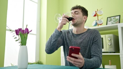 Canvas Print - Young hispanic man drinking glass of wine using smartphone at dinning room