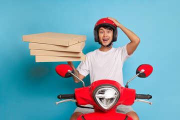 Wall Mural - Happy guy rides on red scooter with pizza boxes in one hand, pressing other hand on his head, fast delivery concept, copy space