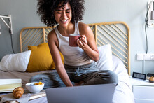 Happy young multiracial latina woman sitting on bed typing on laptop while having morning coffee and eating breakfast.