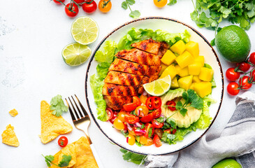 Wall Mural - Yummy salad with grilled chicken fillet with mango, spicy salsa, tomatoes, cilantro, red onion and lettuce in tex-mex style, white table background, top view