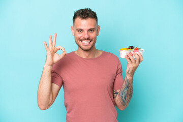 Wall Mural - Young caucasian man holding a bowl of fruit isolated on blue background showing ok sign with fingers