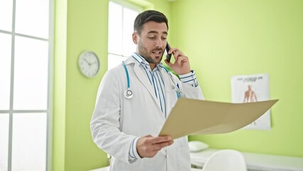 Poster - Young hispanic man doctor reading medical report talking on smartphone at clinic