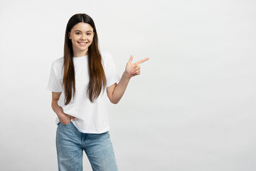 Smiling  teenage girl wearing white t shirt and stylish jeans, pointing finger at copy space, place for text. Happy teen showing to empty place, advertisement. Shopping concept 