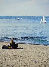Vertical Back View Of People Resting On Sandy Travemuende Beach With Sailboats In The Background