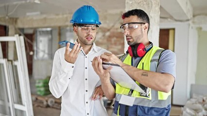 Canvas Print - Two men builders and architect writing on document sending voice message by smartphone at construction site