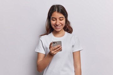 Positive dark haired Near East woman smiles happily checks notification on smartphone messages online dressed in casual t shirt isolated over white background search new friends at social network