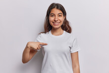 Smiling Dark Haired Young Woman From Iran Demonstrates Her New White Casual T Shirt Being Glad After Making Purchase Indicates At Blank Copy Space For Your Designing Content Or Logo Poses Indoor