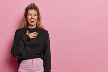 Wall Mural - Horizontal shot of European woman holds hands on chest and smiles from happiness being fascinated dressed in casual black jumper and trousers isolated over pink background copy space for your text