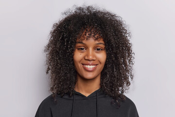 Wall Mural - Portrait of happy curly haired teenage girl smiles pleasantly looks directly at camera dressed in casual black sweatshirt isolated over white background beams with joy has genuine smile on face