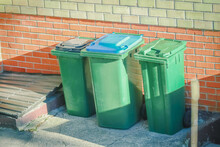 Three Green Garbage Bins Standing Outside The Red Brick Building In Sunlight With Long Dark Shadows
