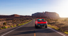 Red Pickup Truck Driving On Scenic Road In The Dry Desert With Red Rocky Mountains In Background