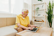 © SHOTPRIME STUDIO - Happy elderly woman with a laptop typing with headphones sitting at home on the couch in a yellow shirt, bright modern interior, lifestyle online communication.