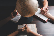Christian Group Praying For Globe And People Around The World On Wooden Table With Bible. Christian Small Group Holding Hands And Praying Together Around A Wooden Table With Bible Page In Homeroom.