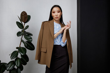 High fashion photo of beautiful elegant young asian woman in pretty brown beige jacket, blazer, blue top, long skirt posing on textured black and white wall, tall flower. Slim figure