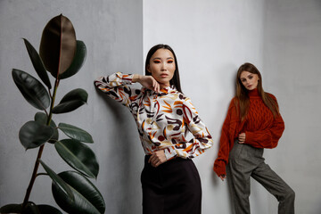 Wall Mural - Two fashion models in orange beige looks, outfits. Sweater, shirt, long black skirt, gray pants, trousers. Beautiful young women.  Asian and european girls posing on textured wall, tall flower