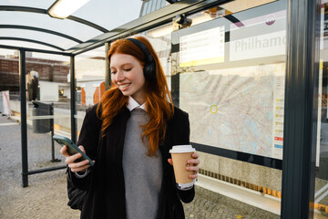 Smiling woman using mobile phone and drinking coffee while standing at bus station