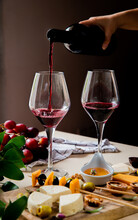 Side View Of Woman Hand Pouring Red Wine Into Glass And Different Kinds Of Cheese Olive Walnut Grape On White Surface And Black Background