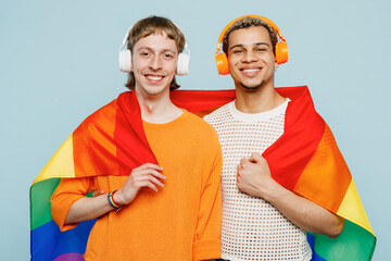 Young happy couple two gay men wear casual clothes wrapped in flag listen muisc in headphones together isolated on pastel plain blue color background studio. Pride day june month love LGBTQ concept.