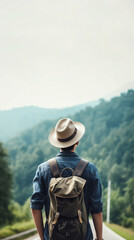 Young man in hat with backpack with his back to the camera standing on an empty open road looking at mountains green valley. Freedom wanderlust adventure traveling concept