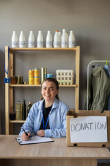 Happy smiling female volunteer with food in box writing on clipboard working at distribution or refugee assistance center. Charity, donation and volunteering