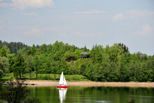 A View Of A Small Boat Or Other Vessel With A Sail Traversing A Vast Yet Shallow River The Coast Of Which Is Surrounded With A Forest, Moor, And A Small Sandy Beach, Seen In Summer In Poland