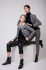 Wall Mural - Two fashion models in gray suit, jacket, blazer, leather pants, black blouse, trousers. Beautiful young woman. Studio shot, portrait. White background. Asian and  blonde. Model sits on cube