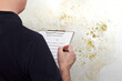 High humidity damage concept: man with a GERMAN inspection checklist in front of a white wall overgrown with mould, mildew or fungus.