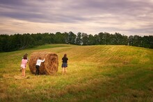 View Of A Group Of Young Girls Attempting To Roll A Round Hay Bale In A Field