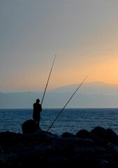 Silhouette of fisherman standing on the shore with rod on sunset sky background