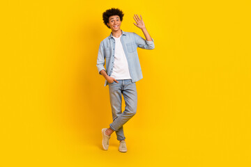 Wall Mural - Full length photo of optimistic cool man dressed denim shirt trousers arm in pocket waving palm say hi isolated on yellow color background