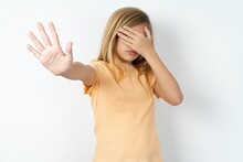 Beautiful Caucasian Teen Girl Wearing Orange T-shirt Over White Wall Covers Eyes With Palm And Doing Stop Gesture, Tries To Hide. Don't Look At Me, I Don't Want To See, Feels Ashamed Or Scared.