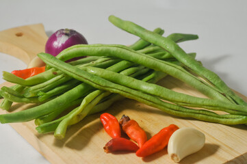 Some green beans, some red chili peppers, two cloves of garlic and a red onion on a wooden chopping board isolated on a white background