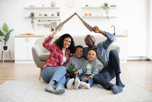 Multicultural Family Holding Cardboard Box Above Heads In Form Of House Roof While Sitting On Carpet In Kitchen. Smiling Adults And Children Updating Moving Day Checklist While Resting On Floor.
