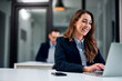 © bnenin - A smiling female employee typing on a laptop with a male colleague in the background.
