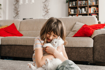 A teenage girl sits on the floor in the living room and plays with her beloved kitten. The child hugs and strokes the pet. Children and cats.