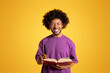 Smiling mature black curly man in purple t-shirt reading book, studying isolated on yellow background