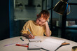 © dikushin - Portrait of tired schoolboy studying at home doing homework sitting at table under light of lamp at evening. Front view of pupil boy writing in exercise book at night in dark with lighting lamp.