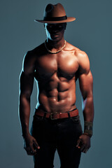Black man, shirtless with strong fashion model and sexy, cow boy style with hat isolated on blue background. Dark, shadow and body with muscular male person, pose with six pack and stylish in studio