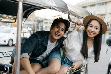 Happy and joyful Young Asian couple traveler tourists riding a tuk tuk tour, rickshaw style transportation on the street in Bangkok in Thailand - people traveling enjoying local culture concept