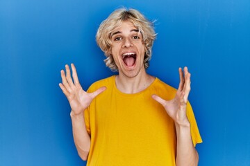 Poster - Young modern man standing over blue background celebrating crazy and amazed for success with arms raised and open eyes screaming excited. winner concept