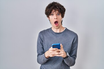 Poster - Young man using smartphone typing a message in shock face, looking skeptical and sarcastic, surprised with open mouth