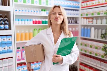 Sticker - Young caucasian woman working at pharmacy drugstore holding box doing inventory looking at the camera blowing a kiss being lovely and sexy. love expression.