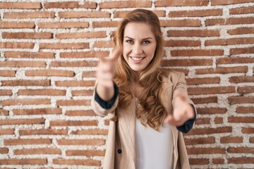 Sticker - Beautiful blonde woman standing over bricks wall looking at the camera smiling with open arms for hug. cheerful expression embracing happiness.