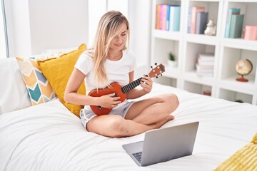 Canvas Print - Young blonde woman having online ukulele lesson sitting on bed at bedroom