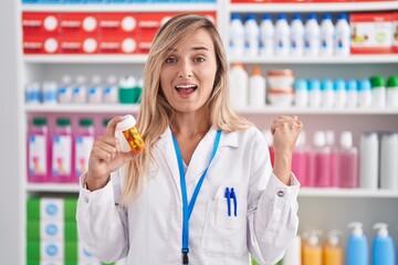 Sticker - Young blonde woman working at pharmacy drugstore holding pills screaming proud, celebrating victory and success very excited with raised arms