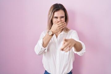 Poster - Young beautiful woman standing over pink background laughing at you, pointing finger to the camera with hand over mouth, shame expression