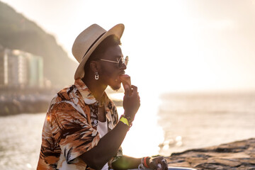 Sunset of a black ethnic man enjoy summer vacation on the beach eating an ice cream with a hat