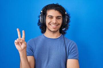 Canvas Print - Hispanic man with curly hair listening to music using headphones smiling with happy face winking at the camera doing victory sign. number two.