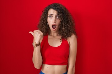 Sticker - Hispanic woman with curly hair standing over red background surprised pointing with hand finger to the side, open mouth amazed expression.